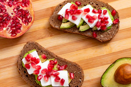 Slice of bread with avocado, cherry tomato and pomegranate seeds, on wood.