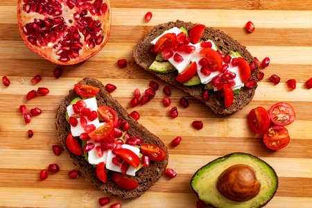 Slice of bread with avocado, cherry tomato and pomegranate seeds, on wood.