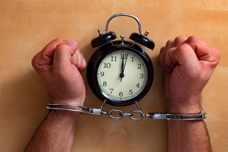 Time in jail. Shackled hands holding an alarm clock on a wooden table.の写真素材