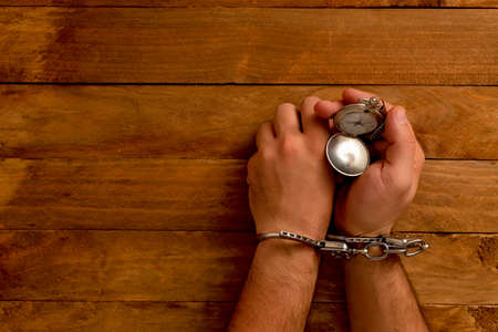 Time in jail. Chained hands holding a hand watch with chain, on a wooden table.の写真素材