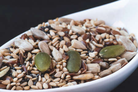 Sesame seeds next to different fruit seeds in a white ceramic spoon. Dark food concept