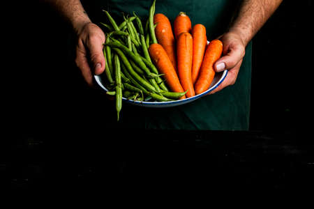 Man with green apron presenting vegetables and garden vegetables. Dark food concept.