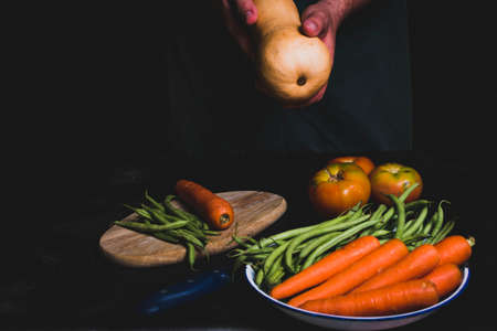 Table full of vegetables with a man in green apron holding a pumpkin with his hands.