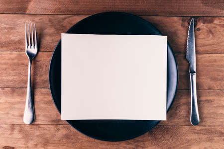 Lunch invitation, consisting of a black plate and cutlery on a wooden table.の写真素材