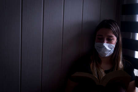 Teenager confined at home by coronavirus COVID-19, reading by the window with a face mask on.の写真素材