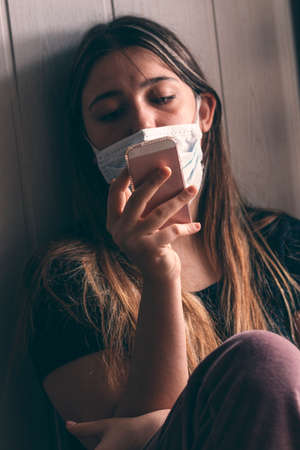 Homebound teenager quarantined with smartphone next to a window with a face mask on.の写真素材
