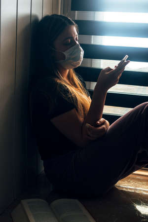Teenager confined at home by coronavirus COVID-19, with the smartphone next to a window with a mask...