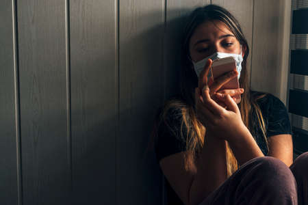 Homebound teenager quarantined with smartphone next to a window with a face mask on.の写真素材