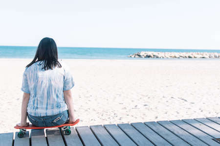 Teen girl in a tie dye shirt sitting on a skateboard looking at the beachの写真素材