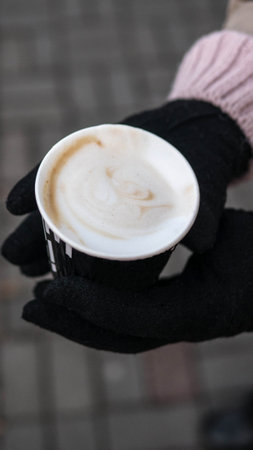 The girl is holding a cappuccino in winter gloves. Fragrant coffee in a glass, a girl on the street...