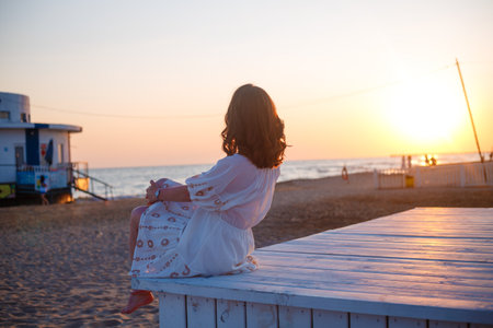 Young woman in white dress sitting on a bench on the beach at sunsetの写真素材