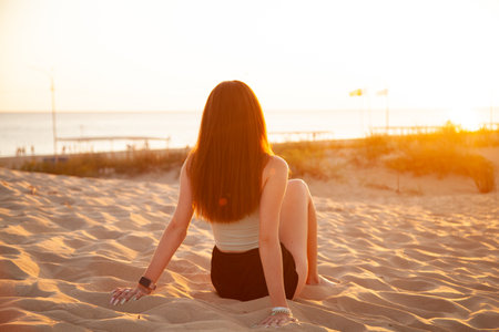 Young woman sitting on the sand in the beach at sunset, back viewの写真素材