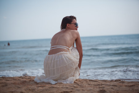 Young woman in white dress and sunglasses sitting on the sand near the seaの写真素材