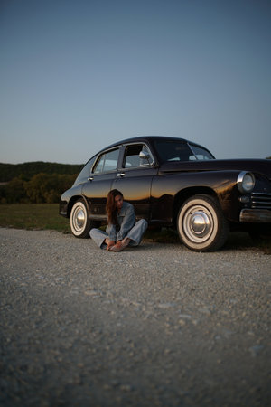 Young woman sitting on the road with her old car in the backgroundの写真素材