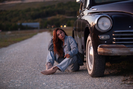 Young woman sitting on the road near the old car at sunset.の写真素材
