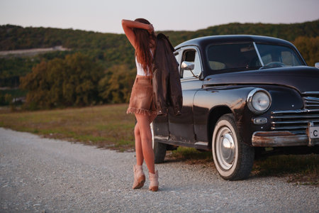 Beautiful young woman standing near old car on the road at sunsetの写真素材