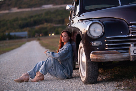 Young stylish woman wearing full denim outfit sitting on a gravel road next to a vintage black carの写真素材