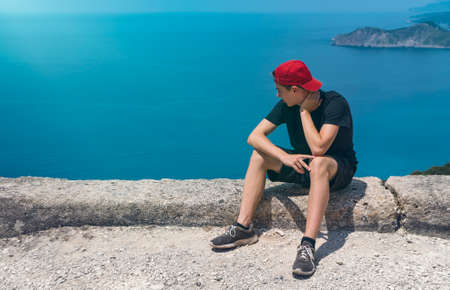 sad young man with baseball cap sits on a wall and looks at the seaの写真素材