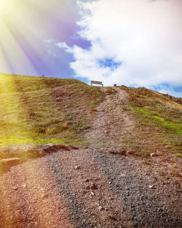 Path in the mountains leads to the old bench in the mountains at the top against blue sky and sun. Vintage image.の写真素材