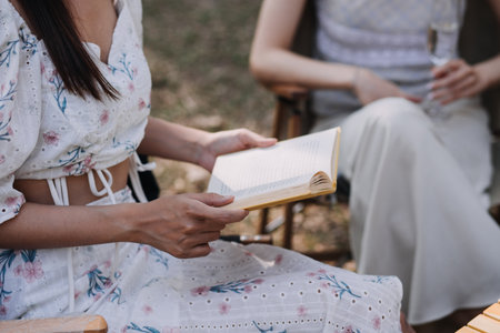 Young pretty brunette twin girls sitting on the grass with legs slightly bent in knees and looking in a brown book, wearing casual coat in autumn sunny weather on blurry background.の写真素材