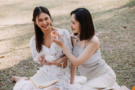 Young pretty brunette twin girls sitting on the grass with legs slightly bent in knees and looking in a brown book, wearing casual coat in autumn sunny weather on blurry background.の写真素材