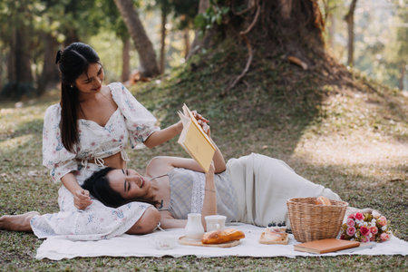 Young pretty brunette twin girls sitting on the grass with legs slightly bent in knees and looking in a brown book, wearing casual coat in autumn sunny weather on blurry background.の写真素材