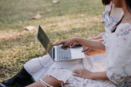 Young pretty brunette twin girls sitting on the grass with legs slightly bent in knees and looking in a brown book, wearing casual coat in autumn sunny weather on blurry background.の写真素材