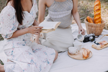 Young pretty brunette twin girls sitting on the grass with legs slightly bent in knees and looking in a brown book, wearing casual coat in autumn sunny weather on blurry background.の写真素材