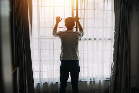Alone man silhouette staring at the window closed with curtains in bedroom. Man stands at window aloneの写真素材