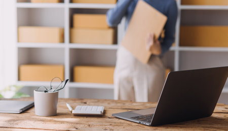 Startup SME small business entrepreneur of freelance Asian woman wearing apron using laptop and box to receive and review orders online to prepare to pack sell to customers, online sme business ideas.の写真素材