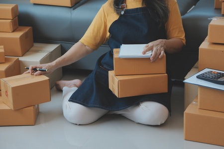 Startup SME small business entrepreneur of freelance Asian woman wearing apron using laptop and box to receive and review orders online to prepare to pack sell to customers, online sme business ideas.の写真素材