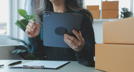 Startup SME small business entrepreneur of freelance Asian woman wearing apron using laptop and box to receive and review orders online to prepare to pack sell to customers, online sme business ideas.の写真素材