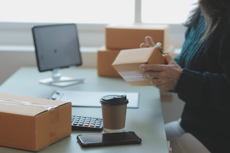 Startup SME small business entrepreneur of freelance Asian woman wearing apron using laptop and box to receive and review orders online to prepare to pack sell to customers, online sme business ideas.の写真素材