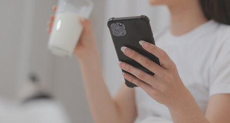 Efficient Young Millennial Girl Sitting on a Bed in the Morning, Uses Laptop Computer and Eats Croissants and Drinks Coffee for Breakfastの写真素材