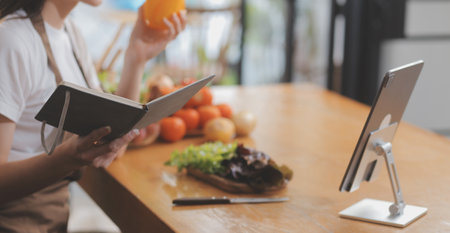 Delicious fruit and vegetables on a table and woman cooking. Housewife is cutting green cucumbers on a wooden board for making fresh salad in the kitchen.の写真素材