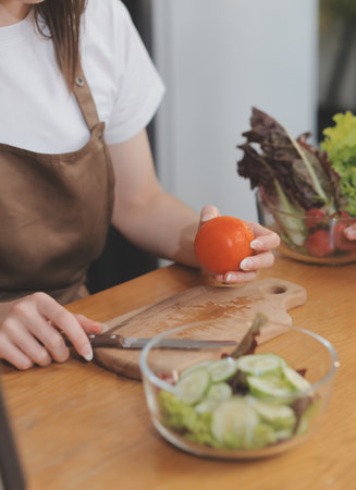 Delicious fruit and vegetables on a table and woman cooking. Housewife is cutting green cucumbers on a wooden board for making fresh salad in the kitchen.の写真素材