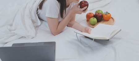 Efficient Young Millennial Girl Sitting on a Bed in the Morning, Uses Laptop Computer and Eats Croissants and Drinks Coffee for Breakfastの写真素材