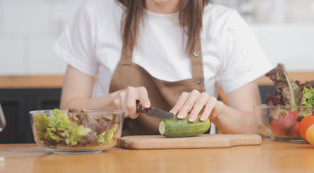Delicious fruit and vegetables on a table and woman cooking. Housewife is cutting green cucumbers on a wooden board for making fresh salad in the kitchen.の写真素材