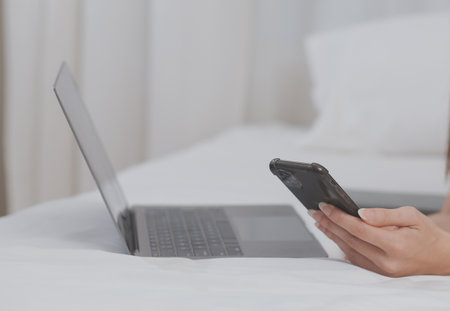 Efficient Young Millennial Girl Sitting on a Bed in the Morning, Uses Laptop Computer and Eats Croissants and Drinks Coffee for Breakfastの写真素材