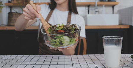 Delicious fruit and vegetables on a table and woman cooking. Housewife is cutting green cucumbers on a wooden board for making fresh salad in the kitchen.の写真素材