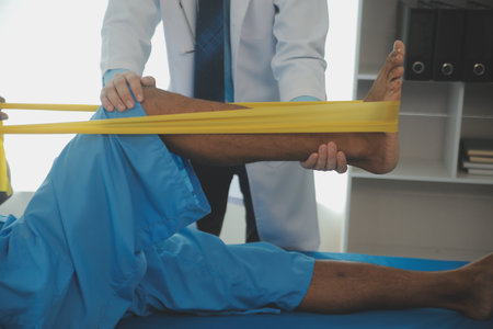 Male patient using elastic band while doing physical exercises for leg muscles at modern clinic,...