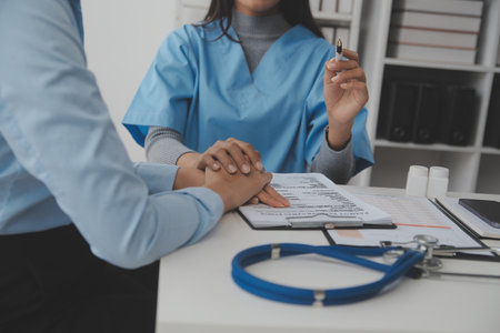 Young woman comes to clinic for heart and lungs checkup. Friendly female doctor sitting at desk in modern medical office, holding stethoscope, listening to patient's breath or heartbeat and smilingの写真素材