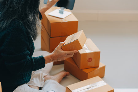 Startup SME small business entrepreneur of freelance Asian woman wearing apron using laptop and box to receive and review orders online to prepare to pack sell to customers, online sme business ideas.の写真素材