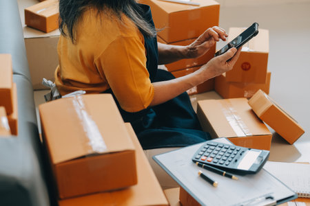 Startup SME small business entrepreneur of freelance Asian woman wearing apron using laptop and box to receive and review orders online to prepare to pack sell to customers, online sme business ideas.の写真素材