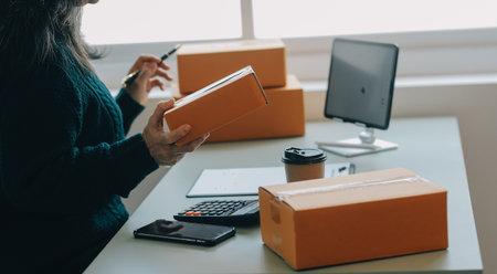 Startup SME small business entrepreneur of freelance Asian woman wearing apron using laptop and box to receive and review orders online to prepare to pack sell to customers, online sme business ideas.の写真素材