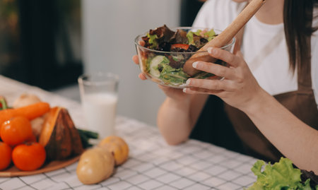 Delicious fruit and vegetables on a table and woman cooking. Housewife is cutting green cucumbers on a wooden board for making fresh salad in the kitchen.の写真素材