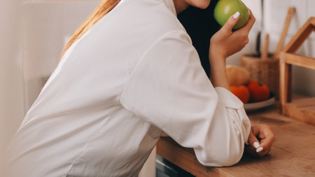 A young woman with a beautiful face in a blue shirt with long hair eating fruit sitting inside the kitchen at home with a laptop and notebook for relaxation, Concept Vacation.の写真素材