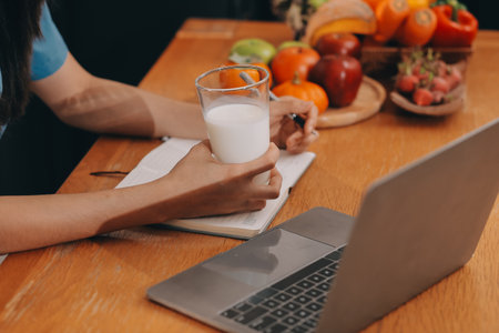 A young woman with a beautiful face in a blue shirt with long hair eating fruit sitting inside the kitchen at home with a laptop and notebook for relaxation, Concept Vacation.の写真素材