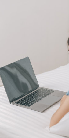 Young Asian woman with beautiful face, long hair, blue shirt lying on bed in white bedroom at home with cup of coffee and video call with laptop talking to relatives on vacation. holiday concept.の写真素材