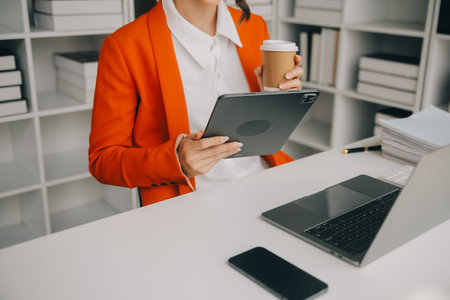 Attractive smiling young asian business woman work at home office, Asian woman working on laptop computer holding tablet.の写真素材